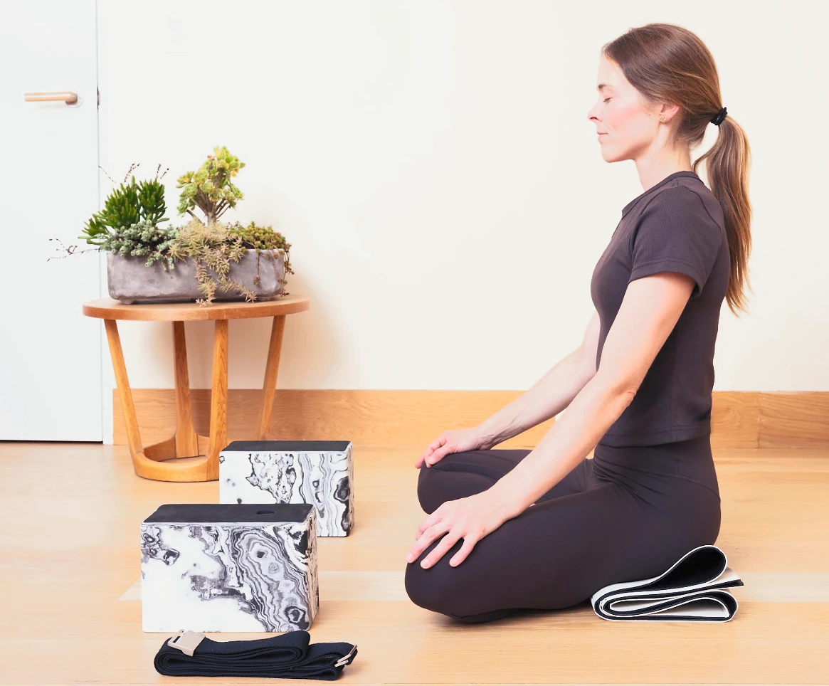 A practitioner seated in meditation on the folded mat, the marble blocks and strap beside her