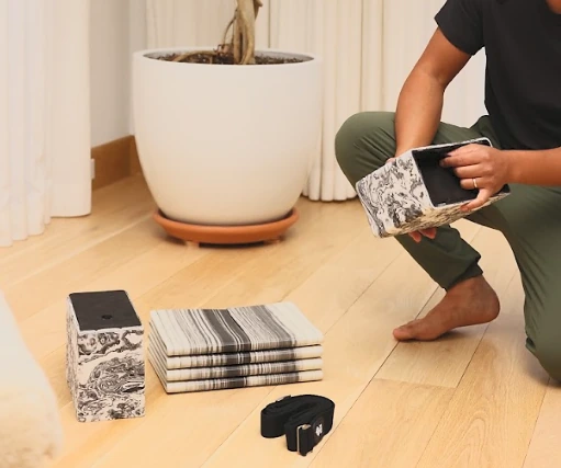 A practitioner holding a hollow Bento Yoga block beside the folded mat, second block, and strap